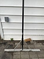 Full view of the wide aluminum landscaping rake against white siding showing the serrated edge and black handle.