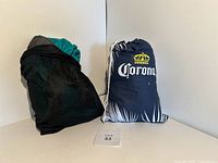 Two packed portable hammocks shown side by side, one in black mesh bag, the other in navy blue Corona drawstring bag with palm design