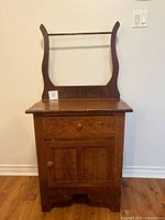 Front view of antique oak washstand showing one drawer with wooden knob, lower cabinet door with knob, and towel bar with curved supports attached at back.