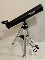 Full side view of the Bushnell Voyager refractor telescope mounted on adjustable aluminum tripod. Shows the telescope tube, tripod with three legs and accessory tray, instruction manual, remote control, eyepieces, dust caps, and pouches laid out below.