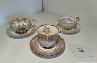 Photo showing three Paragon teacups with matching saucers arranged together: one with pink and gold scrollwork and roses, one with blue cornflowers and gold, and one with pink rose clusters and gold lace trim.