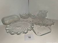 All seven clear glass serving dishes grouped together showing diverse shapes and textures, including ripple swirl, basket-weave, scalloped edges, divided sections, hobnail texture, floral/starburst pattern, and etched details.
