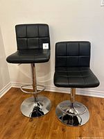 Pair of black faux leather bar stools with chrome pedestal bases side by side showing one stool at maximum height and other at minimum height, highlighting adjustable height feature and cushion upholstery.