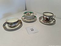 Three vintage bone china teacups and saucers shown together on white background, displaying varying floral and bird designs and gold trim on cups and saucers.