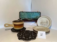 Full lot view showing ceramic pot with lid on black cast iron trivets and four scenic stoneware plates stacked to side with lot number card