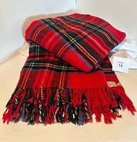 The folded wool tartan throw showing the vibrant classic red with green, navy, yellow, and white plaid pattern and fringe ends.