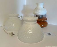 Photo showing two hobnail-style milk glass lamp shades with ruffled edges placed in front of the matching milk glass and faux wood lamp base with metal fitter ring and electrical cord visible.