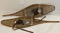 Pair of vintage wooden snowshoes with pointed tails, rawhide laced webbing, brown leather bindings with buckles, laid side by side on floor.