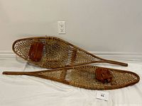 Pair of vintage wooden snowshoes lying flat on a white sheet, showing overall condition, wide rawhide lattice webbing, and tan suede bindings.