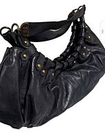 Front view of black leather handbag showing gathered leather design, brass-tone studs and rings, and wide strap with BCBG logo hardware.
