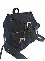 Front view of black canvas and leather backpack showing flap with buckle closure, two front buckle pockets, and adjustable leather strap.
