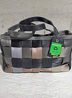 Front view of Harveys woven seatbelt bag showing its checkerboard weave in black, grey, tan, and beige colors, with dual handles and a green Harveys tag attached.