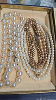 Three pearl necklaces on fabric background inside a box, showing vintage white pearls, gold-tone pearls, and synthetic large beads.