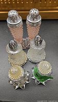 Top view displaying six vintage salt and pepper shakers arranged in three sets on black surface, showcasing metal tops and glass bodies of different designs including pink pressed glass, clear textured glass, and decorative green bird base on one pair.