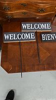 Set of three wooden yard signs placed on wooden furniture showing the 'WELCOME' and 'BIENVENUE' text and metal stakes attached beneath each sign.