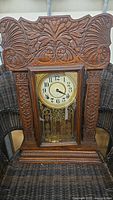 Full front view of the wooden mantle clock showing carved floral and shell designs on the frame and glass door covering the pendulum area.