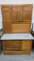 Front view of wooden Hoosier cabinet with metal work surface, multiple upper wooden doors including roll-top tambour door, and lower drawers and cabinet doors.
