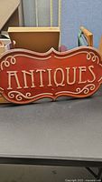 Front view of the red metal sign with white raised letters spelling 'ANTIQUES' and decorative scrollwork on edges.