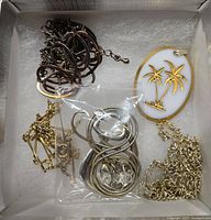 Top-down photo showing assorted chain necklaces and a white enamel vintage compact with gold palm trees.