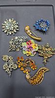 Top-down view of 10 vintage brooches laid out on a black surface showing a variety of floral and abstract designs with rhinestones and enamel colors.