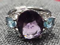 Front view showing oval amethyst center stone flanked by blue topaz stones in silver setting on grey textured background.