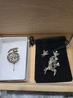Photo showing a silver tone Scottish Thistle brooch with a stick pin in a white box, alongside an Austrian rock pic brooch and matching clip-on earrings placed on a black velvet pouch.