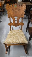 Front view of vintage wooden nursery rocker with intricately carved thistle motif backrest and upholstered seat.