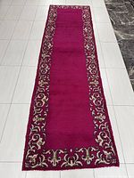 Photo showing full length of the deep red rectangular runner rug with decorative cream/beige floral borders on a white tiled floor.