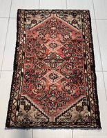 Full view of the entire rug laid flat on a white tiled floor showing the red central medallion and surrounding ivory and black geometric designs.