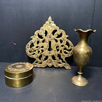 All three antique brass items in the lot displayed against a black background: a vase, lidded container, and decorative door piece.