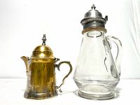 Pair of antique jugs showing the glass syrup jug with pewter lid and silver plated mini creamer jug side by side