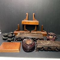 The photo shows a total of 10 wooden display stands grouped together against a black background. The stands include two tall easel type stands, several flat rectangular and round plinths, and multiple intricately carved stands with claw foot designs and ornate detailing in a variety of dark brown wood tones and natural wood finishes.
