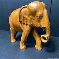 Front and side angled views of large hand carved teak elephant sculpture showing carved details on ears, trunk, and legs.