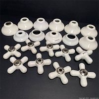 Top-down view showing fourteen porcelain sink knobs with metal centers and fourteen varying porcelain pipe covers of different shapes and sizes arranged on a black surface.