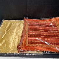 Lot of six silk pillow shams wrapped in plastic, showing one gold tone sham and a stack of orange tone shams with gold beaded grid and embroidery.