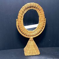 Front view of the oval vanity mirror with wicker frame and base against a plain dark background.
