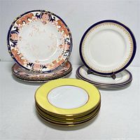 Full lot of 12 plates arranged in three stacks showing Wedgwood Imari pattern, blue and gold Wedgwood plates, and yellow rim plates.
