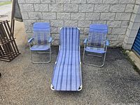 Two light blue metal frame folding patio chairs and one matching lounger with striped fabric seat placed outdoors against a stone wall.