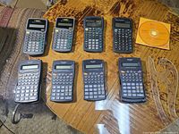 Top view of eight calculators from Texas Instruments and Sharp, plus instructional CD on wooden table.
