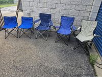 Five fold-up chairs, four blue and one beige, displayed outside against a stone wall and blue door.