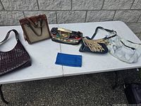 Six vintage purses laid out on a white folding table against a concrete wall, showing variety of styles and colors.
