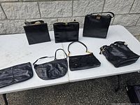Seven black vintage purses on outdoor table showing variety of styles and sizes.