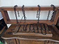 Wooden rectangular ceiling pot holder frame with four attached black metal hanging chains and eight large S-shaped metal hooks laid out on top of wooden antique furniture.