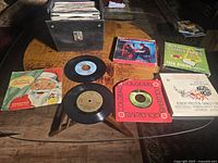 Photo shows black vinyl carry case with metal latch, partially open, containing numerous 45rpm vinyl records and three reel-to-reel tape boxes placed outside the case on a wooden table.