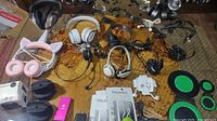 Wide shot on a glass table showing various over-ear headphones, mostly wired, including white, black, and pink cat ear headphones, along with packaging and accessories.
