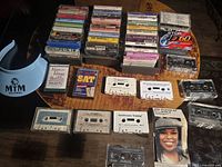 Large group of cassette tapes arranged on wooden surface with MTM Records blue sun visor to left side.
