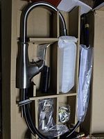 Packaging of faucet components laid out in cardboard box showing brushed nickel faucet body, soap dispenser, and flexible hoses.