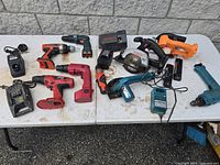Table display of multiple used electric and battery powered handheld tools in various colors including drills, saws, and chargers without batteries.