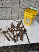 Lot of old hand tools arranged on a white table with a yellow bucket in the background, showing hammers, chisels, and an ax head in used, rusty condition.