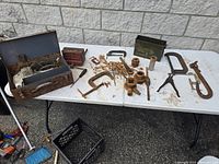 Overview of lot showing rusty old hand tools, clamps, green ammo box bottom, and other rusty parts laid out on white folding table